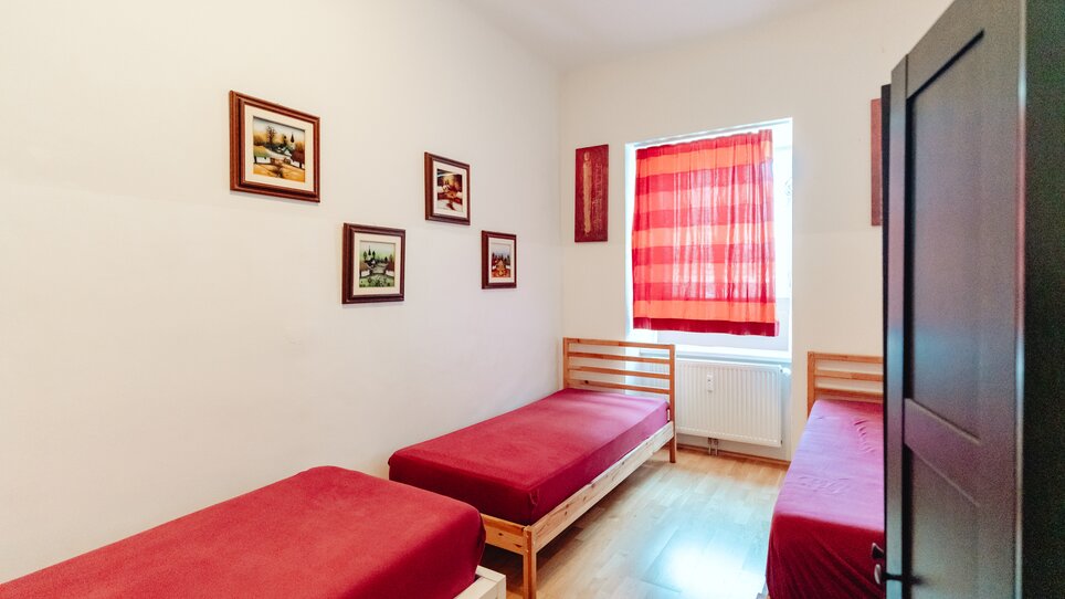 Ein helles Zimmer mit drei Einzelbetten, roter Bettwäsche und Bildern an der Wand. | © Lilians Apartments