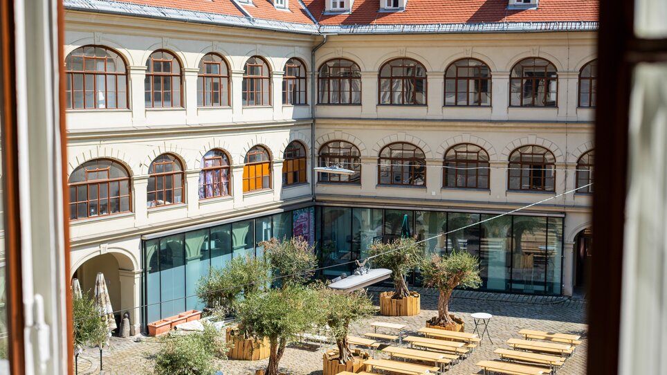 Blick auf einen Innenhof mit Olivenbäumen, Tischen und einer modernen Glasfront. | © HERO HOMES Design Apartments