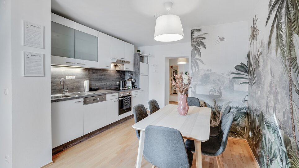 Ein helles Küchen- und Esszimmer mit Pflanzentapete und eleganten Möbeln. | © HERO HOMES Design Apartments