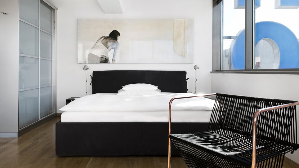 Heller Raum mit Doppelbett, Kunstwerk und einem schwarzen Sessel. | © Augarten Art Hotel