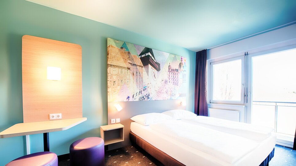 Ein Zimmer im B&B Hotel Graz-Hbf mit Doppelbett, Tisch und Hockern. | © B&B Hotels 2023