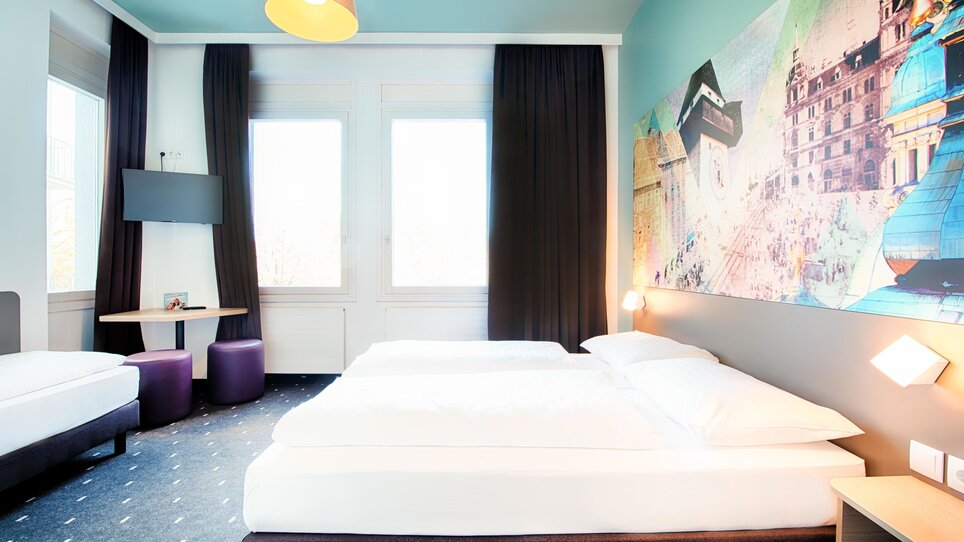 Helles Hotelzimmer mit Doppelbett, einem zusätzlichen Einzelbett und moderner Wandgestaltung. | © B&B Hotels 2023