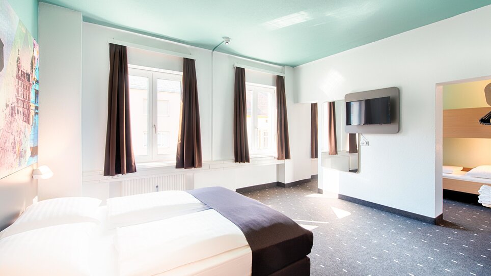 Blick in ein Hotelzimmer im B&B Hotel Graz-Hbf mit Doppelbett, TV und Schreibtisch und einem Durchgang zum Nebenzimmer. | © B&B Hotels 2023