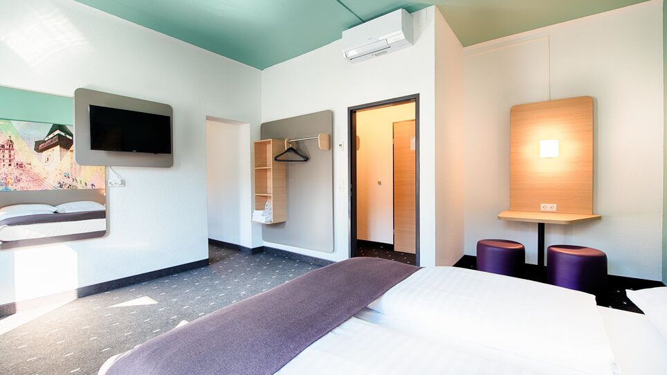 Blick in ein Hotelzimmer im B&B Hotel Graz-Hbf mit Doppelbett, TV und Schreibtisch und einem kleinen Flur zum Nebenzimmer. | © B&B Hotels 2023