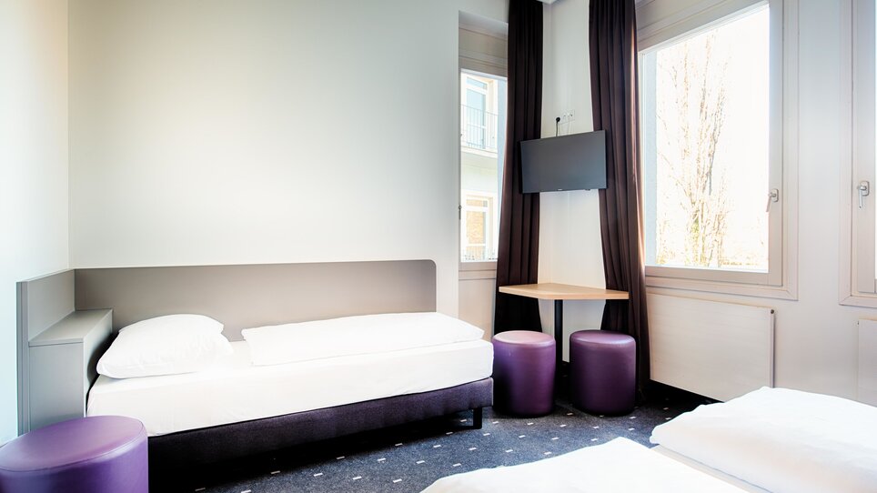 Ein einfaches Hotelzimmer mit drei Betten und einem Tisch. | © B&B Hotels 2023