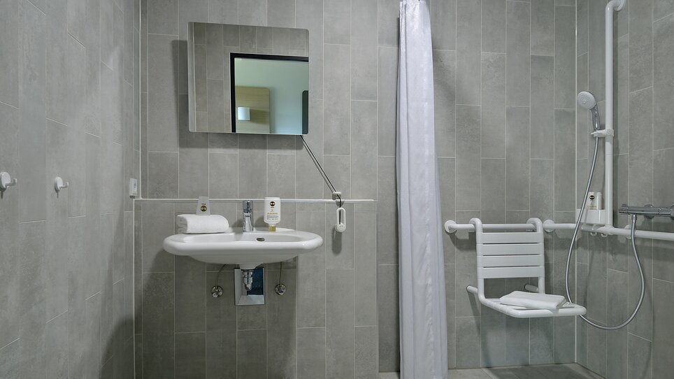 Un bagno accessibile con barre di sostegno e sedia da doccia. | © B&B Hotel Graz City Süd