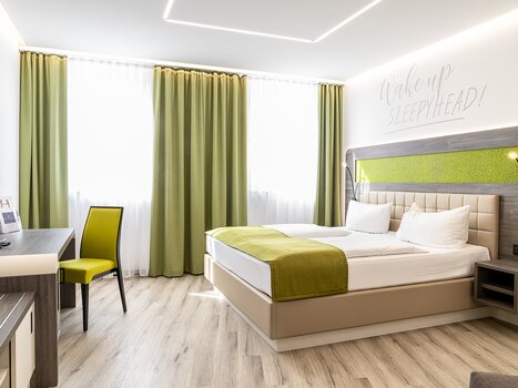 A bedroom with a double bed, TV, and desk. Text on the wall: 'Wake up Sleepyhead!'. | © Chrizz Rooms