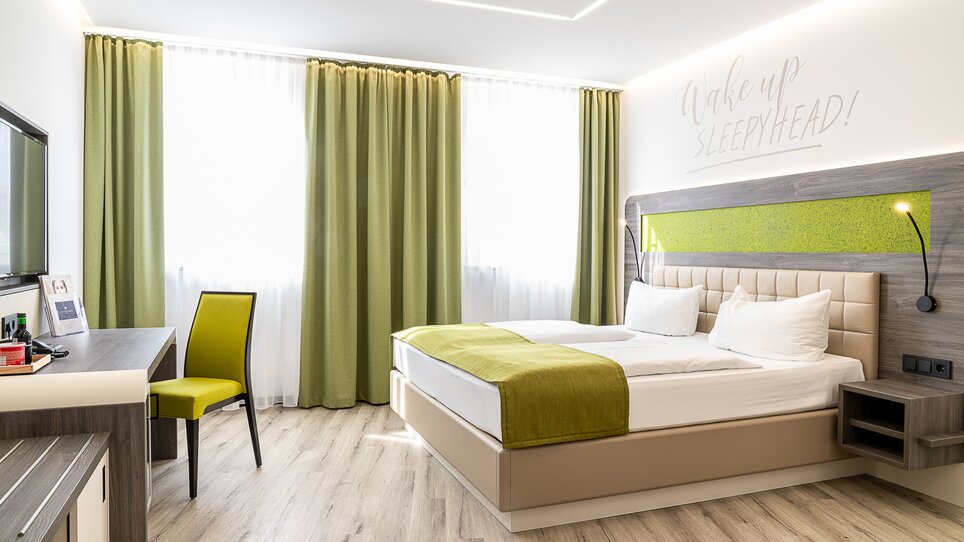 Una camera da letto con un letto matrimoniale, TV e scrivania. Testo sulla parete: 'Wake up Sleepyhead!'. | © Chrizz Rooms