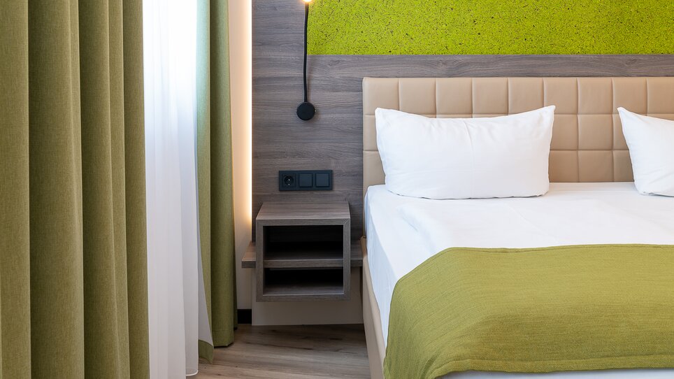 Vista di una camera da letto con letto, comodino e tende in toni di verde. | © Chrizz Rooms