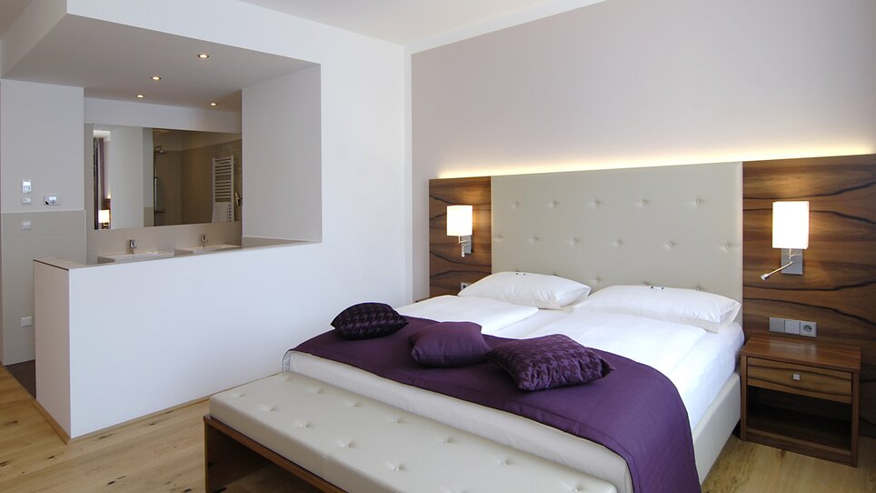 A modern bedroom featuring a double bed with a bathroom area in the background. | © Hotel Feichtinger