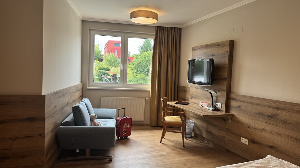 A modern hotel room featuring a desk, TV, and a view through a window onto greenery. | © Der Marienhof