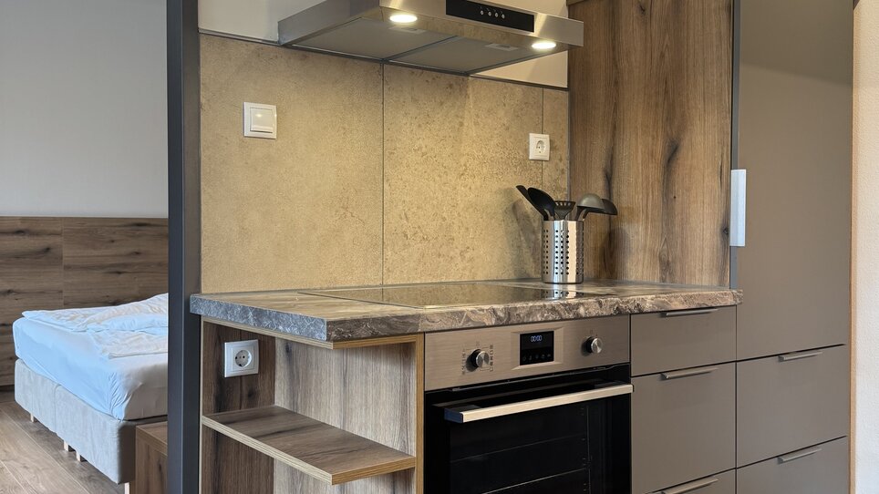 Compact kitchen area featuring an oven, extractor hood, and wood shelving. | © Der Marienhof