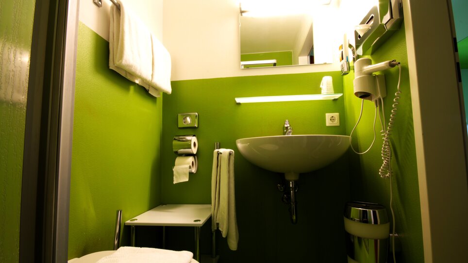 Kompaktes Badezimmer mit grüner Wandfarbe, Waschbecken und Handtüchern. | © Hotel Greenrooms