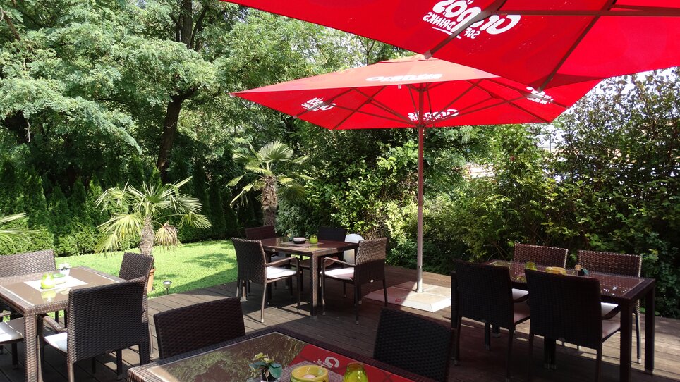 Tische und Stühle unter roten Sonnenschirmen in einem Garten.