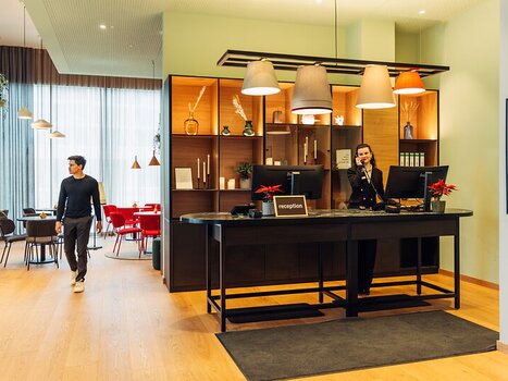 A bright reception area with a staff member on the phone at harry's home Graz-Smart City hotel. | © harry's home Graz-Smart City - Daniel Zangerl