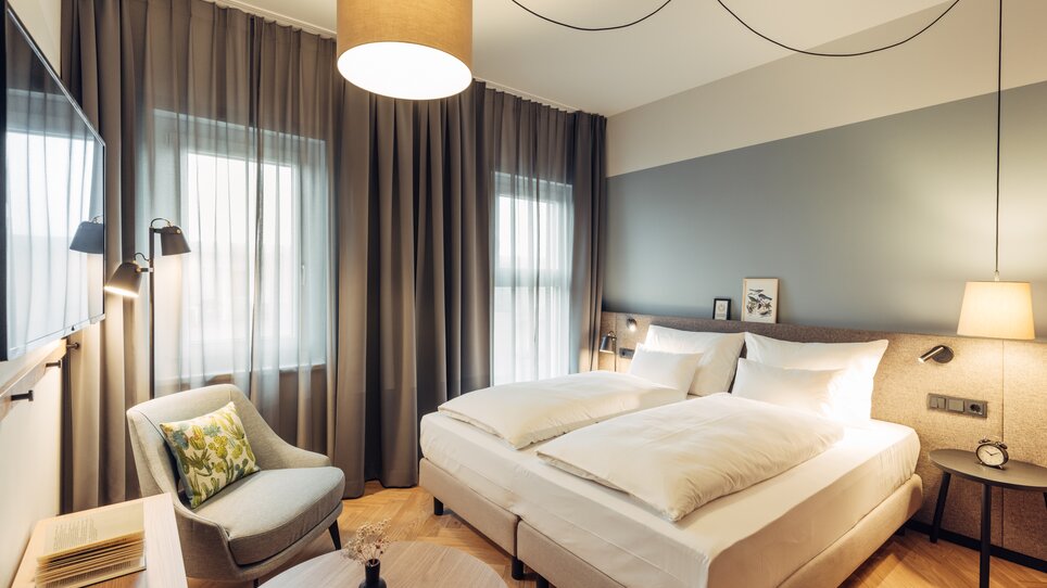 Bright bedroom with two beds, TV and chair at the harry's home Graz-Smart City. | © harry’s home hotels - Daniel Zangerl