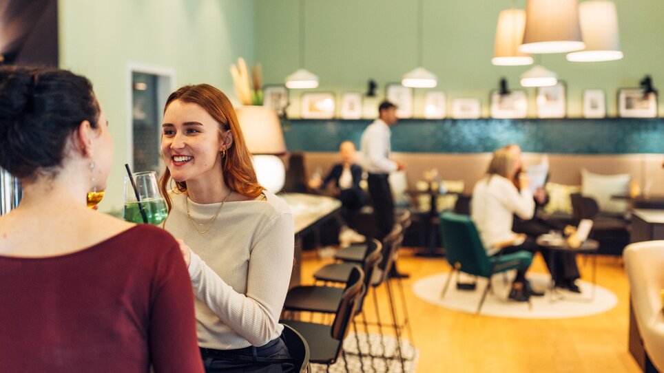 Two women enjoy drinks in a modern bar while other guests relax at the harry's home Graz-Smart City. | © harry’s home hotels - Daniel Zangerl