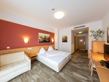 Bright hotel room with a double bed, sofa, and desk. | © Harry Schiffer