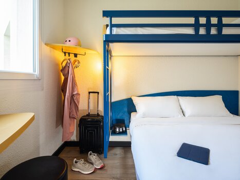 Brightly decorated room featuring a double bed and an elevated bed, suitcase, and clothing. | © Ibis Budget
