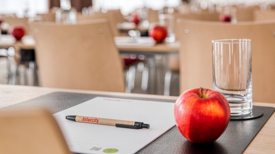 Ein Tisch mit einem Stift, einem Block, einem Glas und einem Apfel in einem Konferenzsaal. | © ICHG