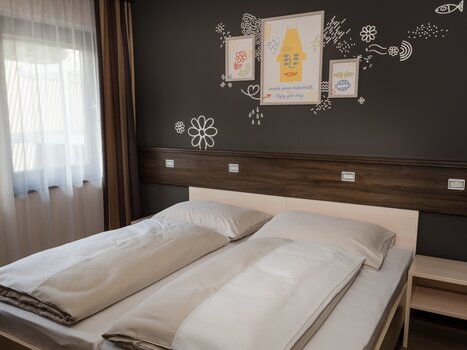 Ein helles Schlafzimmer mit Doppelbett, schlichtem Design und modernen Wanddekorationen. | © Jufa Hotels