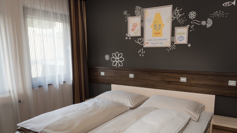 Ein helles Schlafzimmer mit Doppelbett, schlichtem Design und modernen Wanddekorationen. | © Jufa Hotels