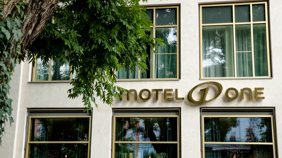 Fassade des Motel One Graz mit großem Schriftzug und Bäumen im Vordergrund. | © Motel One Austria GmbH