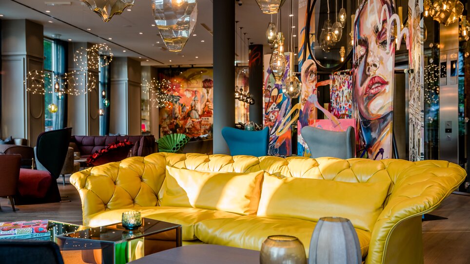 Bequeme gelbe Couch und künstlerische Wandmalerei in einer stylischen Lounge. | © Motel One Austria GmbH