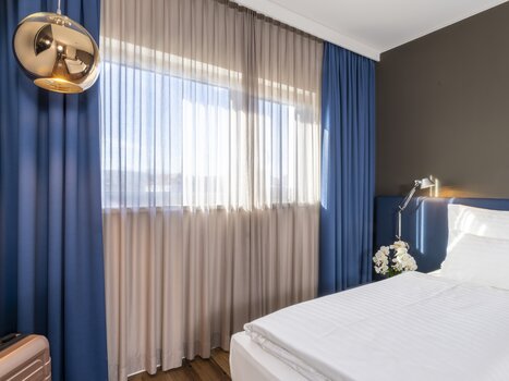 Bright double room with large windows and blue curtains. | © Plaza INN Graz
