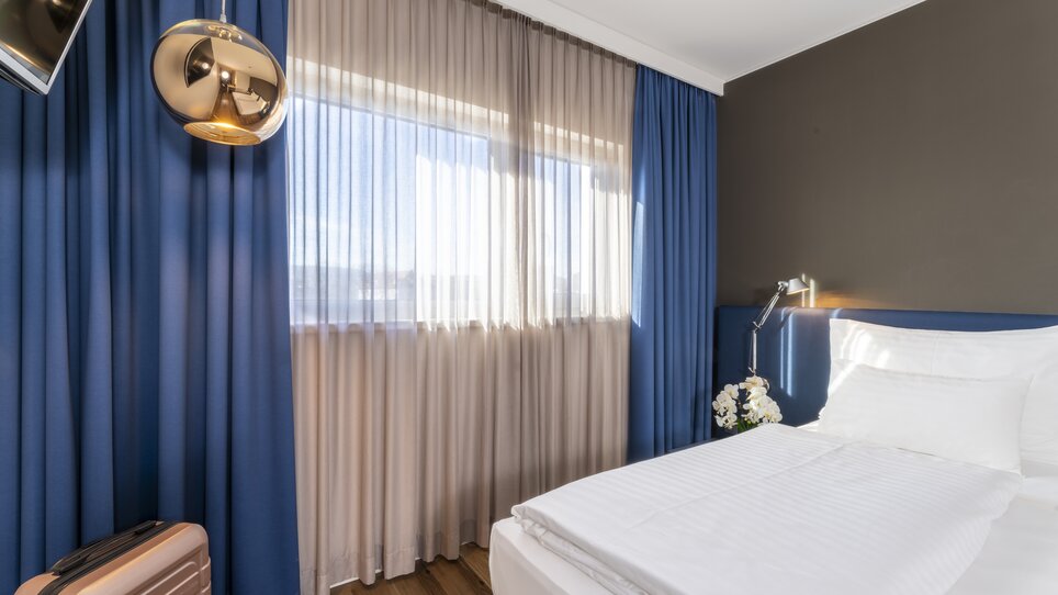 Helles Doppelzimmer mit großen Fenstern und blauen Vorhängen. | © Plaza INN Graz