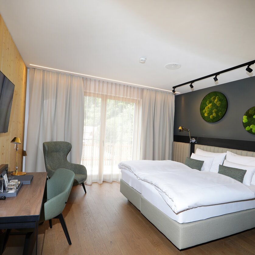 A modern double room decorated in shades of gray, featuring a bed, a desk, a TV, and a large window. Several green decorative elements with moss hang on the wall. | © Hotel Steiermarkhof