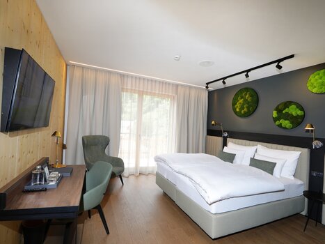 Moderna camera doppia nei toni del grigio, con letto, scrivania, televisore e ampia vetrata. Alle pareti sono appesi diversi elementi decorativi verdi con muschio. | © Hotel Steiermarkhof