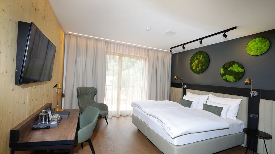A modern double room decorated in shades of gray, featuring a bed, a desk, a TV, and a large window. Several green decorative elements with moss hang on the wall. | © Hotel Steiermarkhof