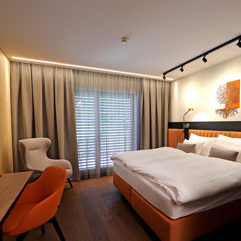 Moderna camera doppia con letto, scrivania, televisore e ampia vetrata. La stanza è arredata con calde tonalità di beige e arancione. | © Hotel Steiermarkhof
