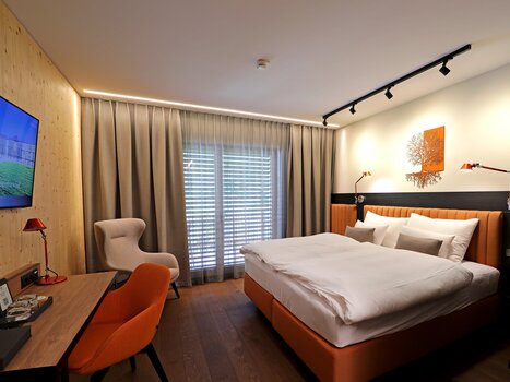 Moderna camera doppia con letto, scrivania, televisore e ampia vetrata. La stanza è arredata con calde tonalità di beige e arancione. | © Hotel Steiermarkhof
