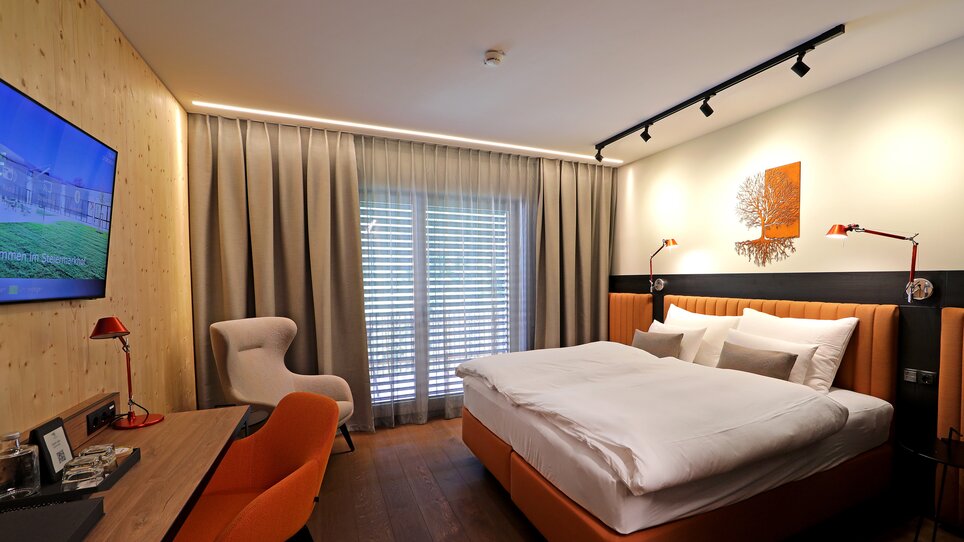A modern double room with a bed, a desk, a television and a large window. The room is decorated in warm shades of beige and orange. | © Hotel Steiermarkhof