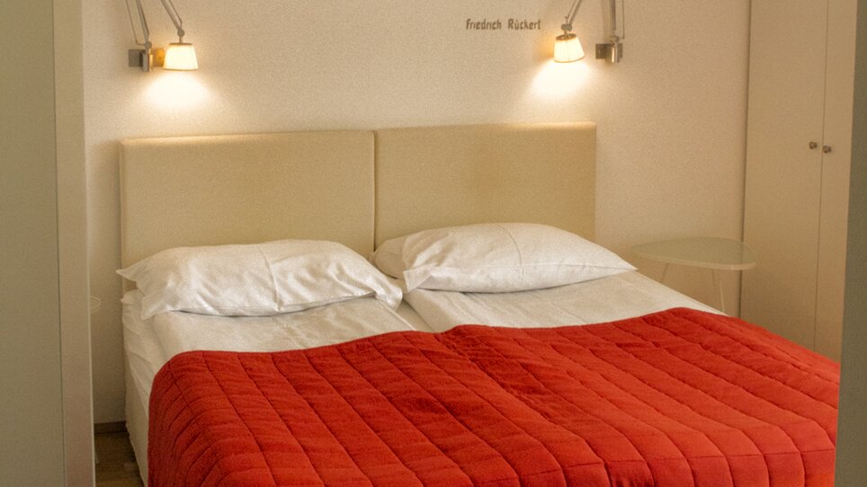 Cozy Bedroom featuring a double bed, a red coverlet, and a wall text by Friedrich Rückert. | © Rueckert
