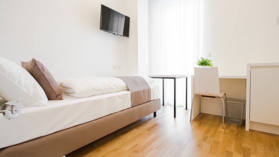Stanza luminosa con letto singolo, scrivania e TV. | © sHome Hotel