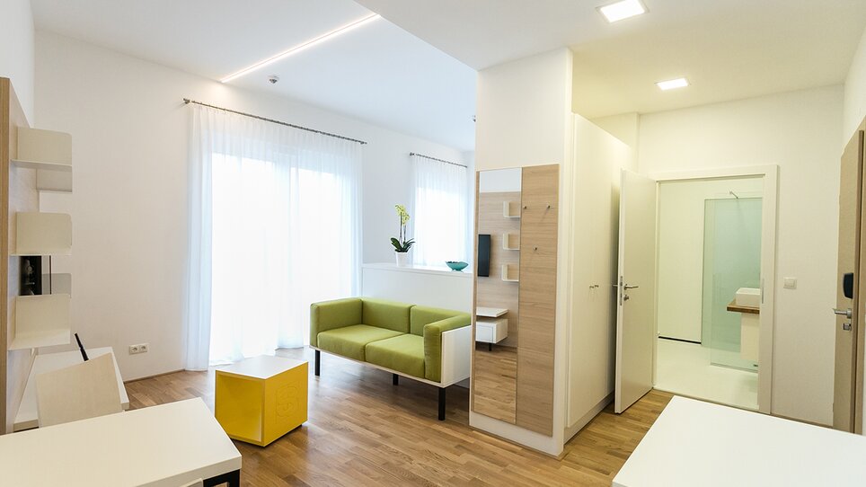 Camera luminosa e moderna con divano verde e tavolo. | © sHome Hotel