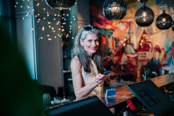 Una donna con i capelli lunghi tiene in mano un cellulare e si trova alla reception dell'hotel. | © Graz Tourismus - Mias Photoart