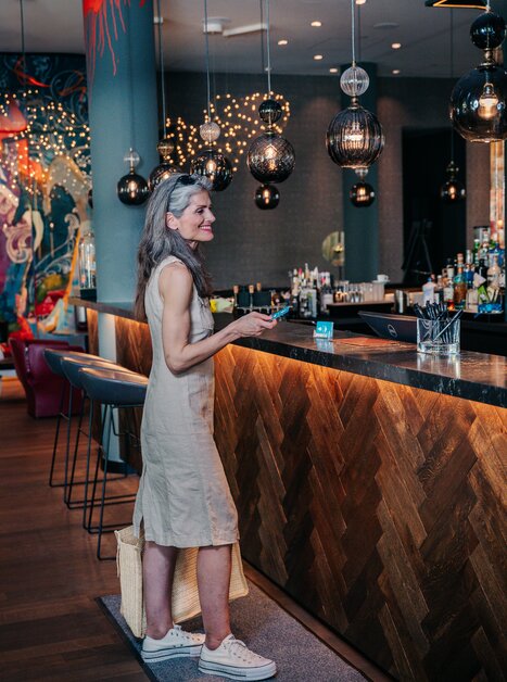 Eine Frau mit grauen Haaren steht an einer Hotel-Rezeption und sieht auf ihr Handy. | © Graz Tourismus - Mias Photoart